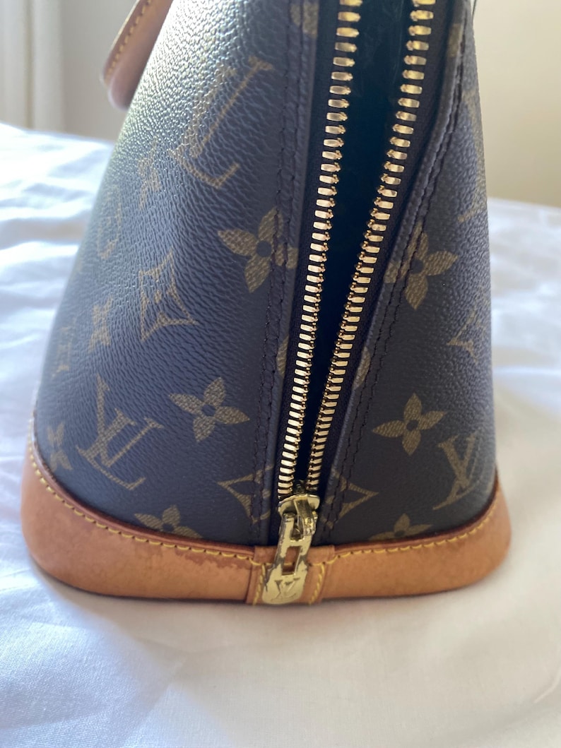 K&ouml;nnte beinhalten: Braune Handtasche mit goldenem Rei&szlig;verschluss und hellbraunem Boden. Die Tasche hat ein sich wiederholendes Muster aus braunen Blumen- und Logodesigns. Der Rei&szlig;verschluss ist ge&ouml;ffnet und gibt den Blick ins Innere frei.