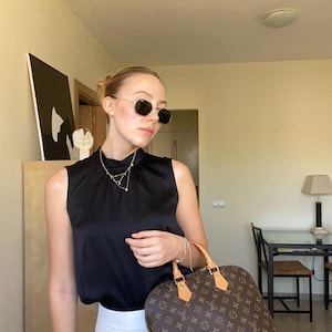 K&ouml;nnte beinhalten: Eine Frau tr&auml;gt eine schwarze Sonnenbrille, ein schwarzes &auml;rmelloses Oberteil und eine wei&szlig;e Hose. Sie h&auml;lt eine braune Handtasche mit Monogrammmuster. Eine goldene Halskette ist sichtbar. Der Hintergrund zeigt einen Raum mit M&ouml;beln und Kunstwerken.