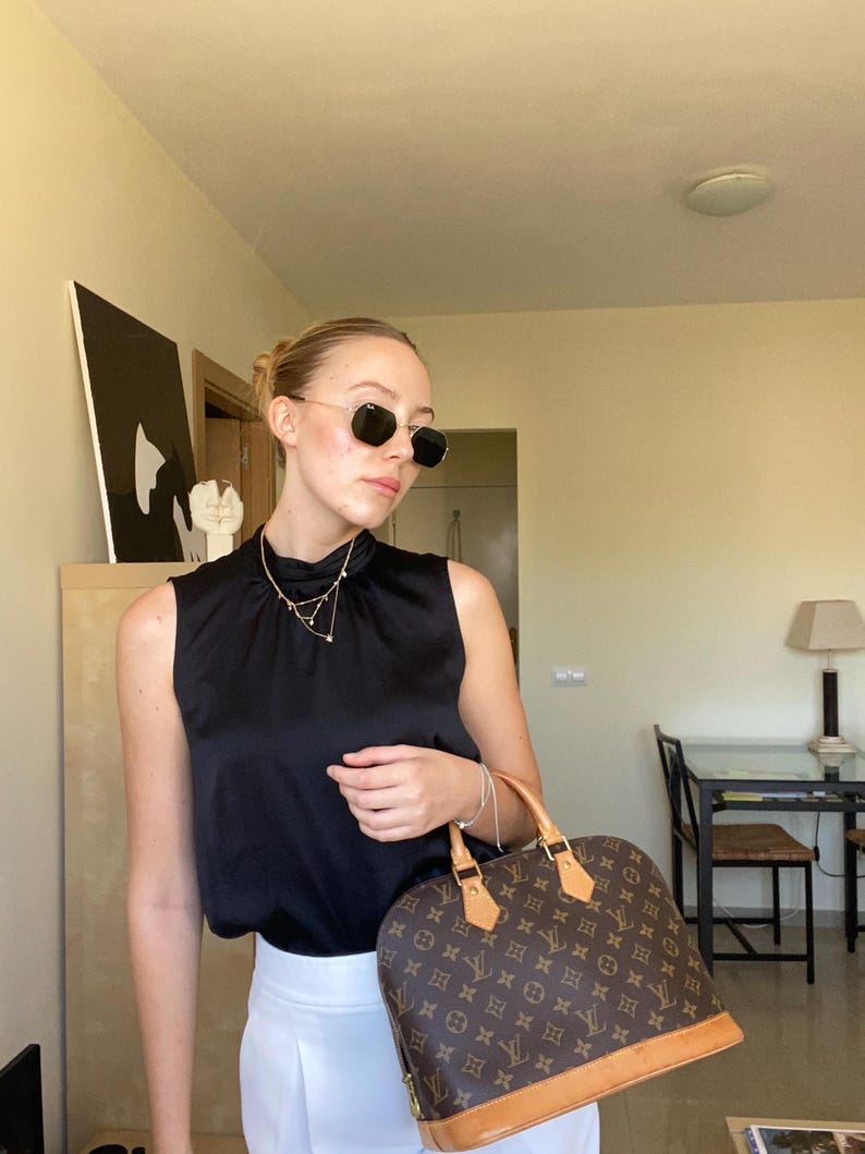 K&ouml;nnte beinhalten: Eine Frau tr&auml;gt eine schwarze Sonnenbrille, ein schwarzes &auml;rmelloses Oberteil und eine wei&szlig;e Hose. Sie h&auml;lt eine braune Handtasche mit Monogrammmuster. Eine goldene Halskette ist sichtbar. Der Hintergrund zeigt einen Raum mit M&ouml;beln und Kunstwerken.