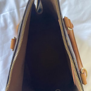K&ouml;nnte beinhalten: Offene Ansicht einer braunen Handtasche mit goldenem Rei&szlig;verschluss und hellbraunen Lederakzenten. Der Innenraum ist dunkelbraun. Die Tasche hat eine strukturierte Form und einen oberen Griff. Die Tasche besteht aus einem gemusterten Material.