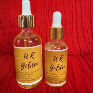 Op de afbeelding: Twee glazen druppelflessen met goudkleurige doppen en witte druppelaars. De flessen hebben gouden etiketten met de tekst "UR Golden" en ingrediëntenlijsten. De flessen zijn gevuld met een heldere vloeistof en gouden vlokken.