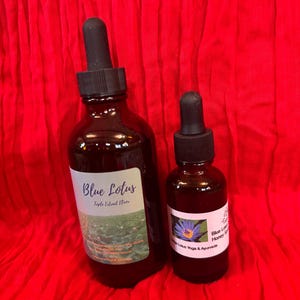 以下が含まれることがあります： 黒いスポイトが付いた2つの茶色のガラス瓶。大きなボトルには「Blue Lotus Triple Extract Elixir」と書かれたラベルが付いています。小さなボトルには、青い蓮の花と「Blue Lotus Honey Syrup」というテキストのラベルが付いています。