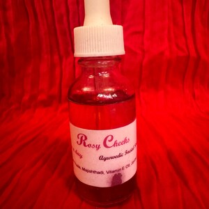 May include: A clear glass bottle with a white dropper top, filled with a red liquid. The bottle has a white label with the text "Rosy Cheeks Ayurvedic Facial Serum". The background is a red fabric.