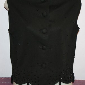 May include: A black vest with a round neckline and a row of four buttons down the front. The vest is embellished with small, scattered beads and a scalloped hemline with small tassels. The vest is displayed on a mannequin.