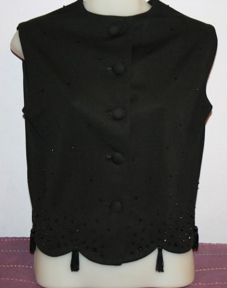 May include: A black vest with a round neckline and a row of four buttons down the front. The vest is embellished with small, scattered beads and a scalloped hemline with small tassels. The vest is displayed on a mannequin.