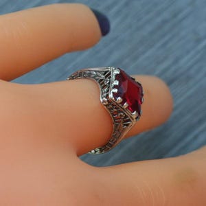 May include: A silver filigree ring with a large, rectangular, deep red gemstone. The ring is on a finger, with a blue background. The ring's intricate design and vibrant gemstone create a striking contrast.