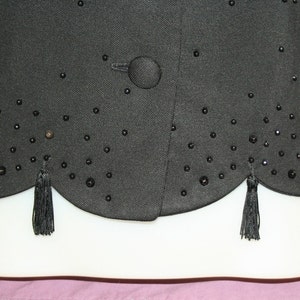 May include: Black vest with a scalloped hemline and small black beaded embellishments. The vest has a single button closure and decorative tassels at the hem. The fabric appears to be a textured weave.