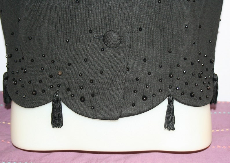 May include: Black vest with a scalloped hemline and small black beaded embellishments. The vest has a single button closure and decorative tassels at the hem. The fabric appears to be a textured weave.