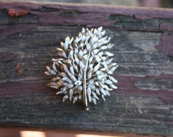 May include: Silver-toned leaf-shaped brooch. The brooch features a detailed design of layered leaves, with a central stem and a pin closure on the back. The brooch is set against a weathered wooden surface.