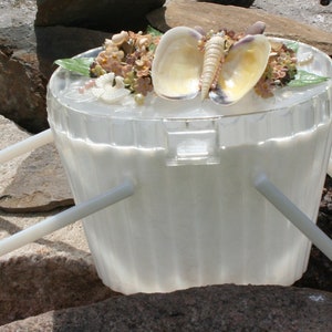 May include: A clear, white plastic bucket with a handle, decorated with seashells and small flowers. The bucket is sitting on a rock surface. The bucket has a lid and a handle.