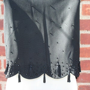 May include: Black sleeveless top with a scalloped hemline and decorative tassels. The top is embellished with small, dark beads scattered across the fabric. The design features a subtle, tailored silhouette, suitable for formal or semi-formal occasions.