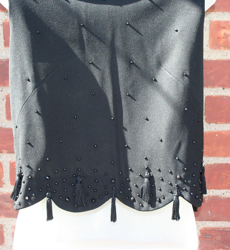 May include: Black sleeveless top with a scalloped hemline and decorative tassels. The top is embellished with small, dark beads scattered across the fabric. The design features a subtle, tailored silhouette, suitable for formal or semi-formal occasions.