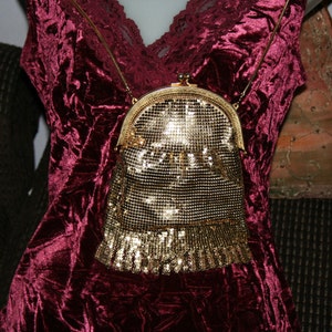 May include: A vintage gold mesh purse with a gold chain strap. The purse has a rounded top and a fringe detail at the bottom. The purse is set against a burgundy velvet background.