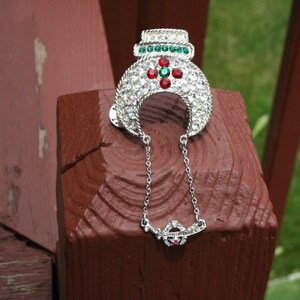 May include: A silver-toned brooch with a decorative design. The brooch features clear rhinestones, with accents of green and red gemstones. A delicate chain with a small pendant hangs from the brooch. The brooch is set against a brown wooden background.