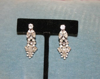 May include: A pair of silver-toned, dangling earrings. Each earring features a cluster of clear, sparkling crystals in a cascading design. The earrings are displayed on a black velvet stand against a blue background.