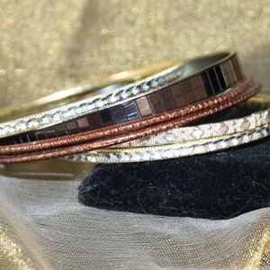 May include: A collection of bangles in various metallic finishes. The bracelets include gold, silver, copper, and a mosaic-style black band. The bangles are stacked and displayed against a gold and black background.
