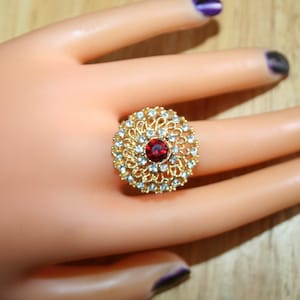 May include: A gold-toned ring with a central red gemstone, surrounded by a halo of smaller clear stones. The ring has an ornate, filigree design. The ring is displayed on a mannequin hand.