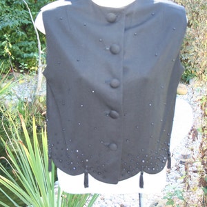 May include: A black vest with a high collar and four large buttons down the front. The vest is decorated with small, scattered embellishments and a scalloped hemline with small tassels. The vest is displayed on a white form.