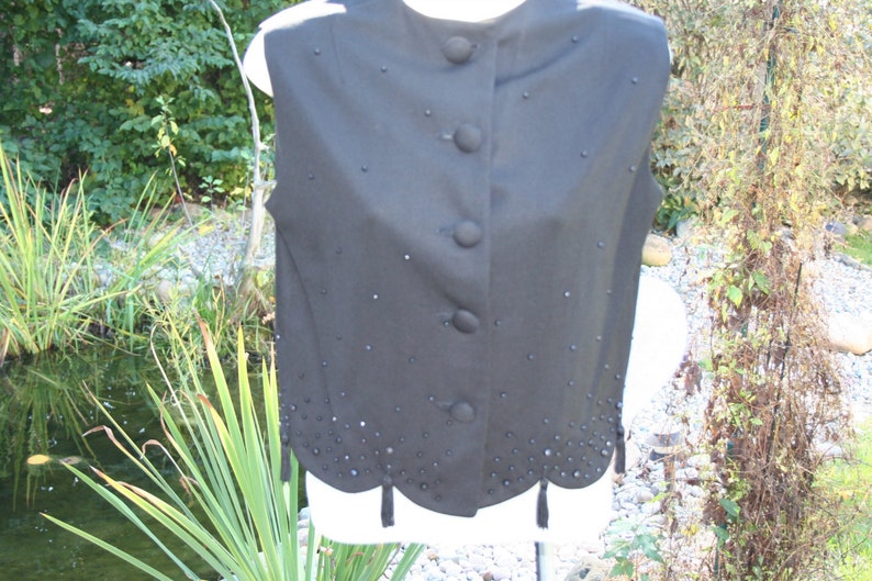 May include: A black vest with a high collar and four large buttons down the front. The vest is decorated with small, scattered embellishments and a scalloped hemline with small tassels. The vest is displayed on a white form.