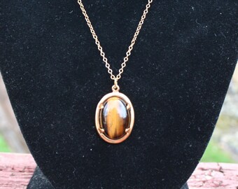 May include: A gold-colored necklace with an oval pendant. The pendant features a brown and gold tiger's eye stone set in a gold-tone frame. The necklace is displayed on a black velvet stand, showcasing the jewelry's design and color.
