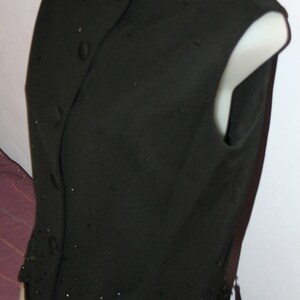 May include: A black sleeveless vest with a round neckline and button closure. The vest is adorned with small, scattered embellishments and a scalloped hemline with small tassels. The garment is displayed on a white mannequin.
