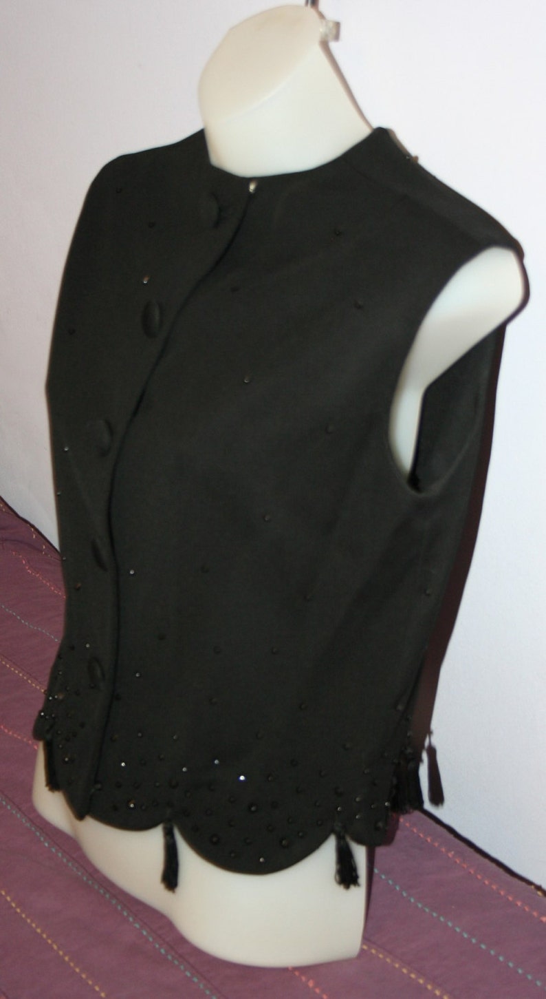 May include: A black sleeveless vest with a round neckline and button closure. The vest is adorned with small, scattered embellishments and a scalloped hemline with small tassels. The garment is displayed on a white mannequin.