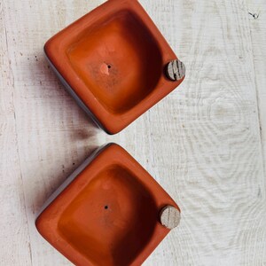 May include: Two terracotta-colored square planters with a small, round, wooden plug on the side. The planters have a small drainage hole in the center. The planters are set on a light-colored wooden surface.