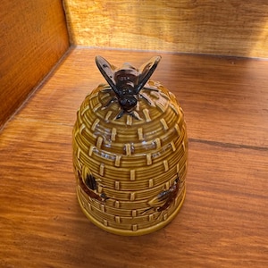 May include: A ceramic honey pot shaped like a beehive, with a dark brown bee figurine on top. The pot is a light brown color with a woven basket design. The pot has two brown bee illustrations on the side.
