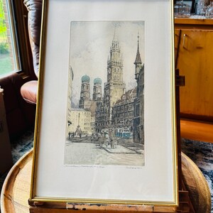 May include: Framed print of a European city street scene, featuring buildings with spires and domes. The print is in a gold frame and displayed on a wooden easel. The easel has the text "AARON BROTHERS" on the top.