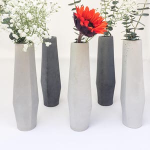 May include: Five gray concrete vases of varying heights with a geometric design. The vases are filled with white flowers and a single orange sunflower.