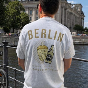 Könnte beinhalten: Ein hellgraues T-Shirt mit dem Wort BERLIN in goldenen Buchstaben oben. Darunter zeigt eine Grafik einen vertikalen Döner-Spieß und eine Bierflasche. Der Text lautet No Doner No Berlin und DÖNER-MODE.