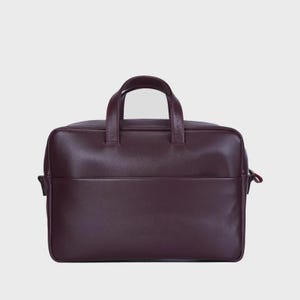 Könnte beinhalten: Eine burgunderfarbene Lederaktentasche mit einem oberen Griff und einer Vordertasche. Die Tasche hat eine rechteckige Form und einen Reißverschluss. Die Aktentasche ist für den Transport von Dokumenten und anderen wichtigen Dingen konzipiert.