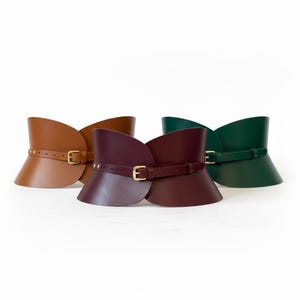 May include: Three wide corset-style belts in tan, burgundy, and dark green leather. Each belt features a smooth, curved silhouette, a thin adjustable strap across the front, and a gold-toned metal buckle with matching hardware accents.