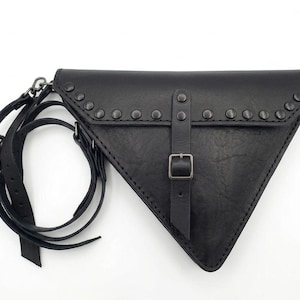 Pode incluir: Bolsa triangular de couro preto com fecho de aba, adornada com tachas prateadas nas bordas. Possui fecho de fivela e alças ajustáveis. Design único e arrojado.