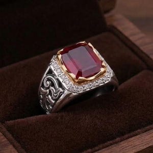 May include: A silver ring featuring a large, rectangular, deep red gemstone set in a gold-toned bezel. The band is decorated with intricate scrollwork engravings and a border of small, clear stones surrounding the central red gemstone.