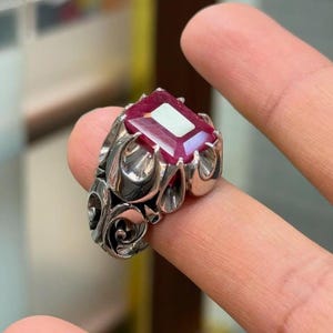 以下が含まれることがあります： 大きな長方形の深い赤色の宝石が爪留めされたシルバーリング。リングのバンド部分は、宝石の台座から側面にかけて、複雑な渦巻き状の有機的な金属パターンが施されています。