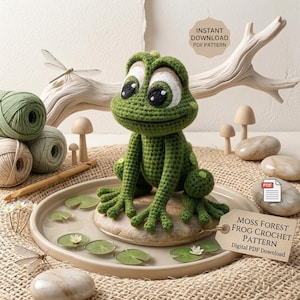 May include: A handmade green crochet frog sits on a stone, surrounded by lily pads and yarn. The frog has large eyes and is part of a crochet pattern. The image includes the text "Instant Download PDF Pattern" and "Moss Forest Frog Crochet Pattern".