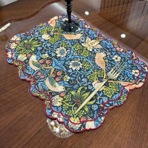 Può includere: Un sottobicchiere decorativo con bordo smerlato, con un motivo floreale e di uccelli dettagliato nei toni del blu, verde, giallo e arancione. Una forchetta dorata poggia sul sottobicchiere, accanto a un bicchiere da vino.