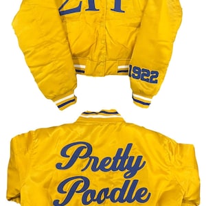 Peut inclure: Veste de bombardier en satin jaune avec lettrage et bordures bleues. Le devant affiche les lettres grecques ΣΓΡ et l'année 1922. Le dos de la veste porte l'inscription "Pretty Poodle" en écriture cursive.