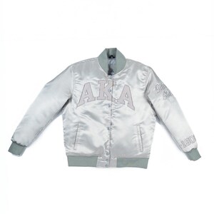 Puede incluir: Chaqueta bomber de satén plateado con detalles en gris claro. La chaqueta presenta las letras "AKA" en gris claro en la parte delantera y texto en las mangas. Tiene cierre de botones y puños y cuello acanalados.