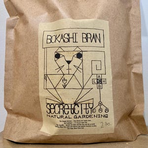 May include: A brown paper bag with a cream label displaying "BOKASHI BRAN" and a geometric cat illustration. The label also states "SECRET CITY NATURAL GARDENING" and lists ingredients. The bag contains 907 grams of product.
