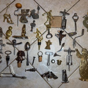 May include: A collection of vintage wine bottle openers and stoppers in various designs, including figures, animals, and abstract shapes. The openers are made of metal and cork, with a variety of colors and finishes. The collection is displayed on a tiled surface.