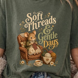 May include: An olive green t-shirt featuring a graphic of a mouse sitting in a brown armchair knitting. The text reads Soft Threads & Gentle Days. Beside the chair is a basket filled with yarn balls and a small teacup on a saucer with floral accents.