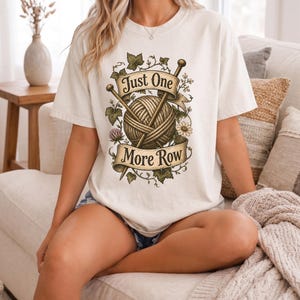 Peut inclure: T-shirt cr&egrave;me avec un motif de fil, d'aiguilles &agrave; tricoter et le texte "Just One More Row". Le motif est entour&eacute; d'&eacute;l&eacute;ments floraux et de feuilles. Le t-shirt est de coupe ample.