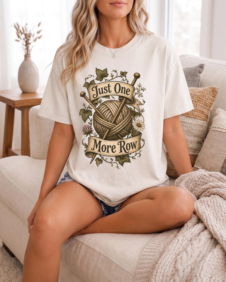 Peut inclure: T-shirt cr&egrave;me avec un motif de fil, d'aiguilles &agrave; tricoter et le texte "Just One More Row". Le motif est entour&eacute; d'&eacute;l&eacute;ments floraux et de feuilles. Le t-shirt est de coupe ample.