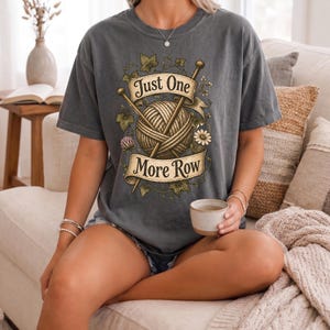 Peut inclure: T-shirt gris fonc&eacute; avec une pelote de laine, des aiguilles &agrave; tricoter et le texte "Just One More Row". Le motif comprend des &eacute;l&eacute;ments floraux et des feuilles. Le t-shirt est de coupe ample.