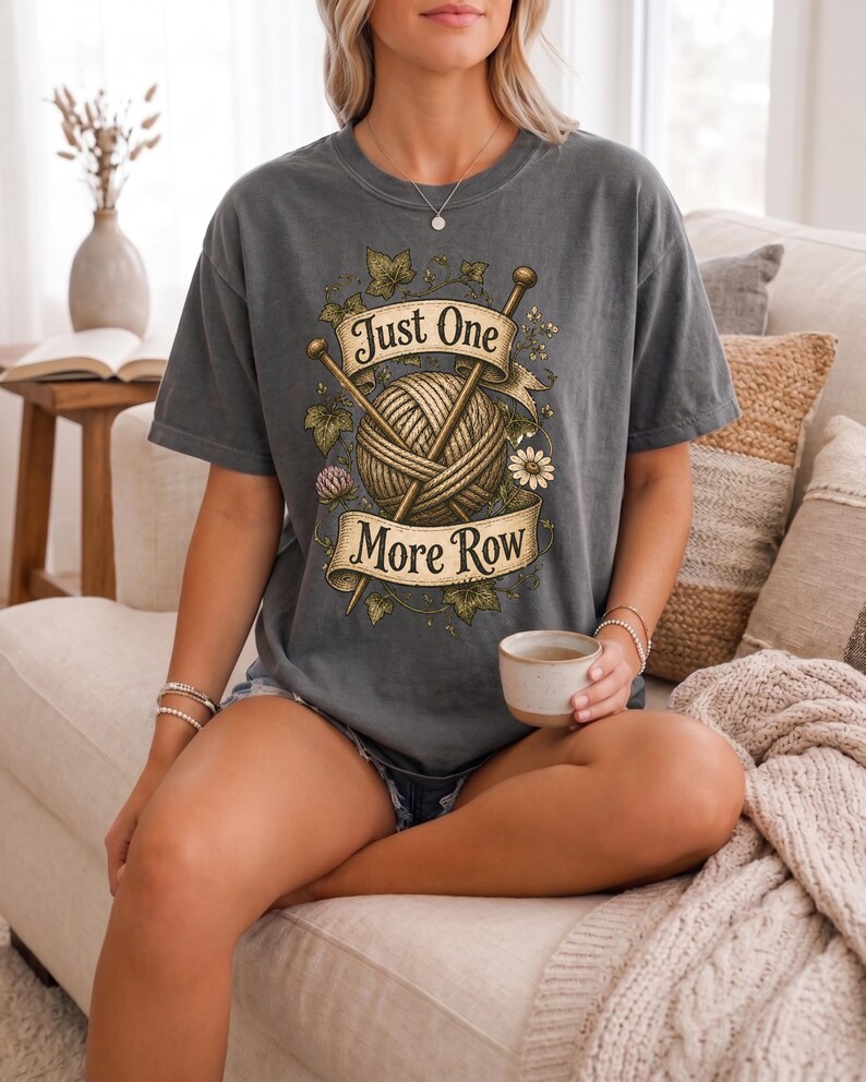 Peut inclure: T-shirt gris fonc&eacute; avec une pelote de laine, des aiguilles &agrave; tricoter et le texte "Just One More Row". Le motif comprend des &eacute;l&eacute;ments floraux et des feuilles. Le t-shirt est de coupe ample.