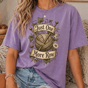 Peut inclure: Un t-shirt violet avec un motif d'une pelote de laine, d'aiguilles &agrave; tricoter et le texte "Just One More Row". Le motif est encadr&eacute; d'&eacute;l&eacute;ments floraux et de feuilles. Le t-shirt est de couleur violet uni.