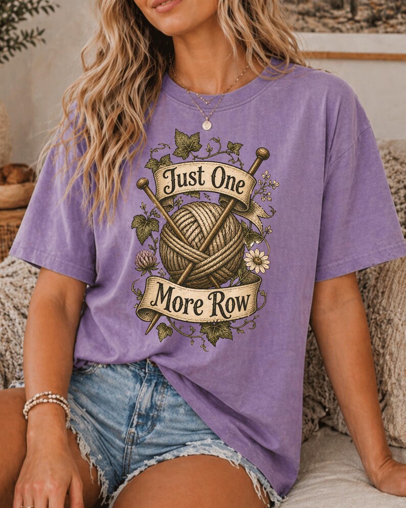 Peut inclure: Un t-shirt violet avec un motif d'une pelote de laine, d'aiguilles &agrave; tricoter et le texte "Just One More Row". Le motif est encadr&eacute; d'&eacute;l&eacute;ments floraux et de feuilles. Le t-shirt est de couleur violet uni.