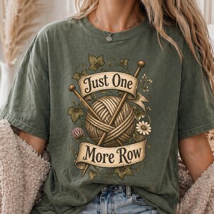 Peut inclure: T-shirt vert olive avec un motif de tricot. Le motif comprend une pelote de laine, des aiguilles &agrave; tricoter et le texte "Just One More Row". Des d&eacute;tails floraux et de feuilles compl&egrave;tent le look inspir&eacute; de l'artisanat.